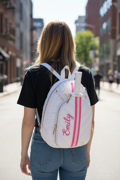 A woman viewed from behind wearing a white quilted backpack with pink stripes and the name 'Emily' in pink script, walking down a city street.