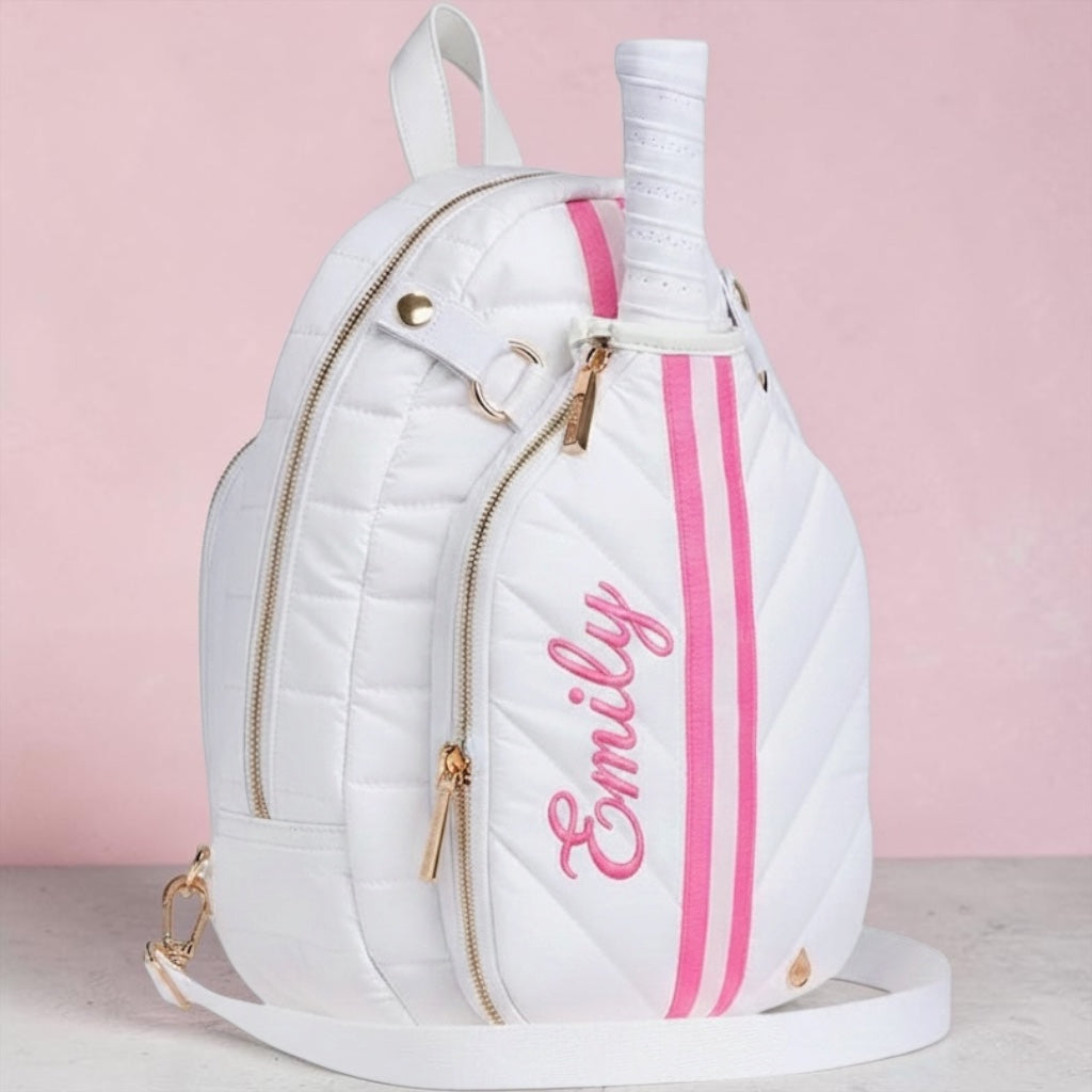 A white quilted backpack designed like a tennis racket head, featuring pink vertical stripes, pink cursive 'Emily' embroidery, and a tennis racket handle tucked into the top slot. The bag has gold-tone hardware and is set against a soft pink background.