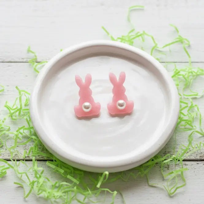 Pink bunny-shaped earrings on a white dish with green Easter grass on a light wooden background