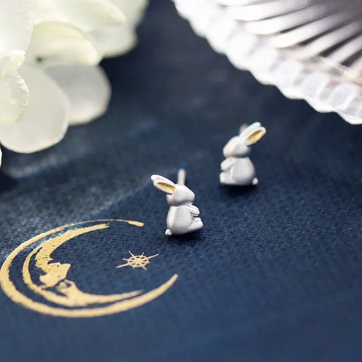 Small rabbit-shaped earrings on a dark blue fabric background with a gold crescent moon design.