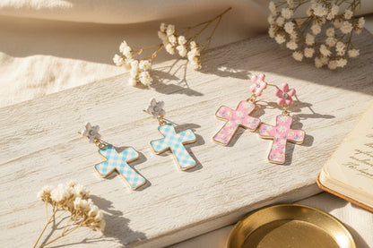 Pair of cross-shaped earrings with checkered patterns on a wooden surface with dried flowers.