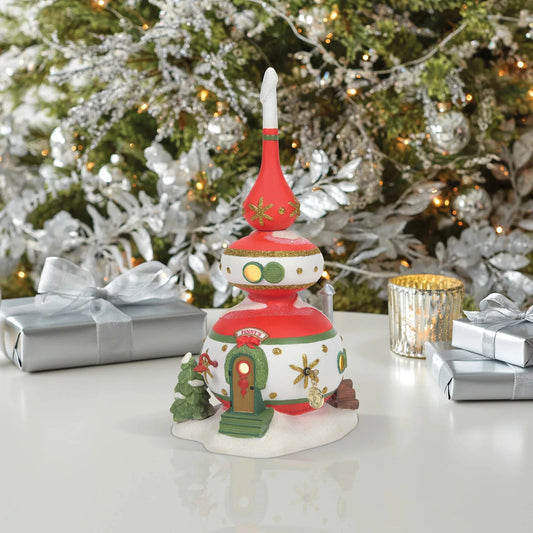 Decorative Christmas village figure with a tree and presents on a table in front of a decorated Christmas tree.