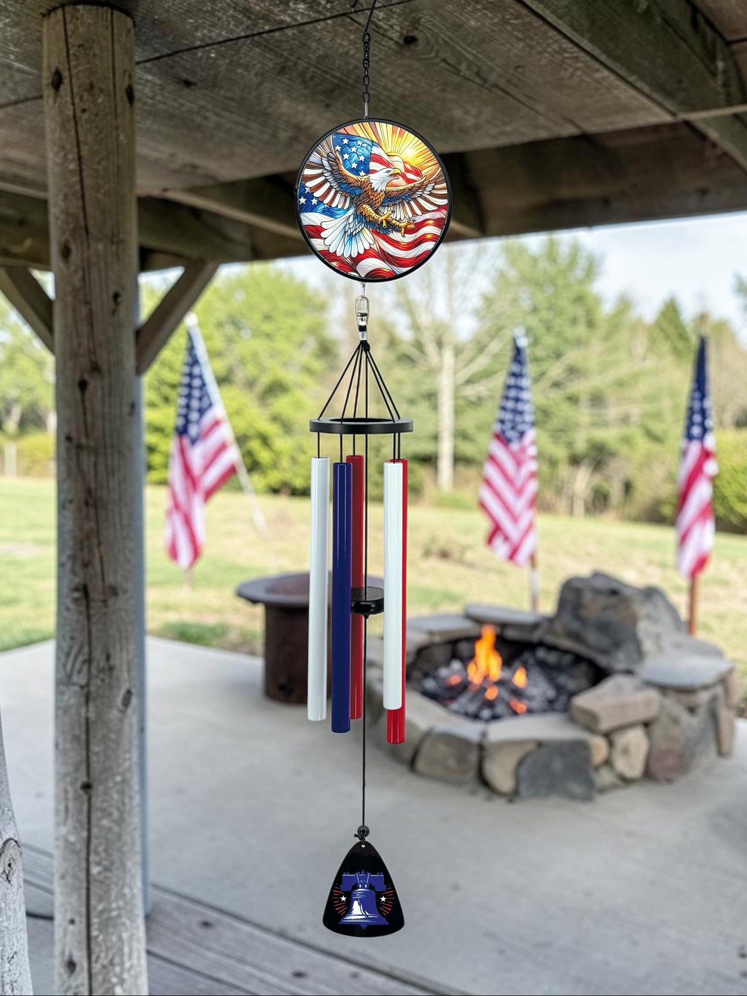 Decorative wind chime with American flag design hanging from a wooden pergola with a fire pit and flags in the background.