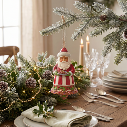 A handcrafted ceramic Santa Claus holiday ornament hangs from a pine branch at a styled holiday table with candles, plates, and greenery.