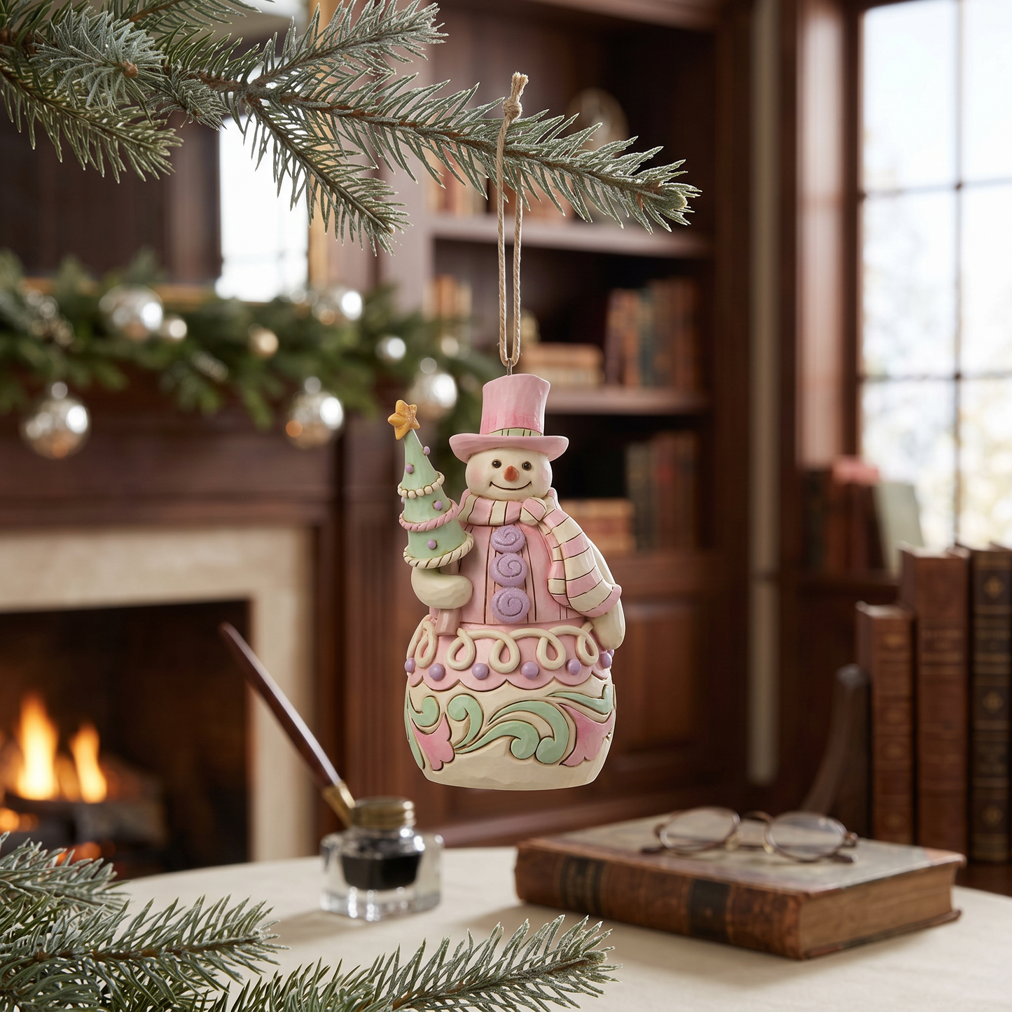 A decorative hanging snowman ornament with a small tree, shown hanging from a pine branch in a cozy indoor holiday setting with a fireplace, books, and glasses in the background.