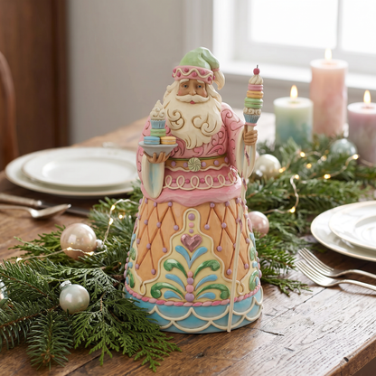 A decorative ceramic Santa Claus figurine dressed in a pink and cream winter outfit, holding a stack of presents in one hand and a rainbow-topped wand in the other, displayed on a rustic wooden table with holiday greenery, plates, candles, and fairy lights.