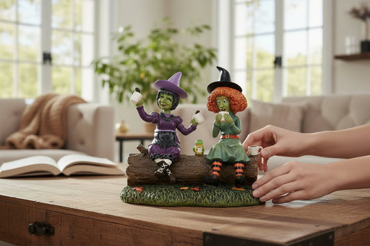 Two witch figurines on a log with a hand reaching towards them, set against a home interior background.