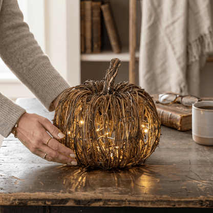 Decorative wicker pumpkin with string lights on a wooden table, held by a person.