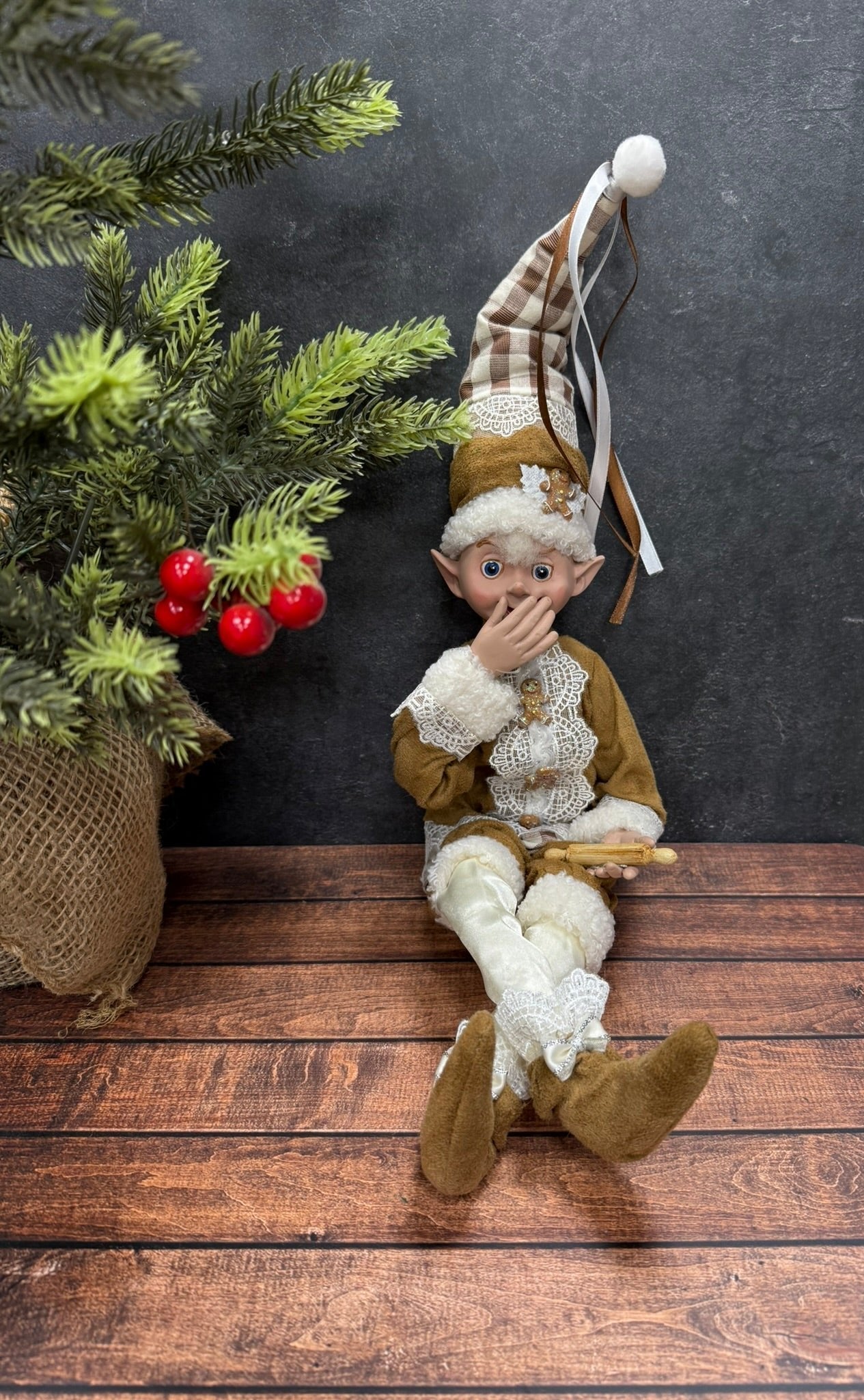 Elf figurine sitting on a wooden surface with a dark background and Christmas tree holding a rolling pin