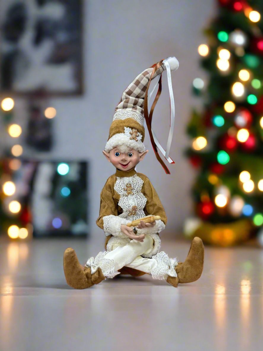 Elf figurine sitting, holding a rolling pin, in front of a decorated Christmas tree.
