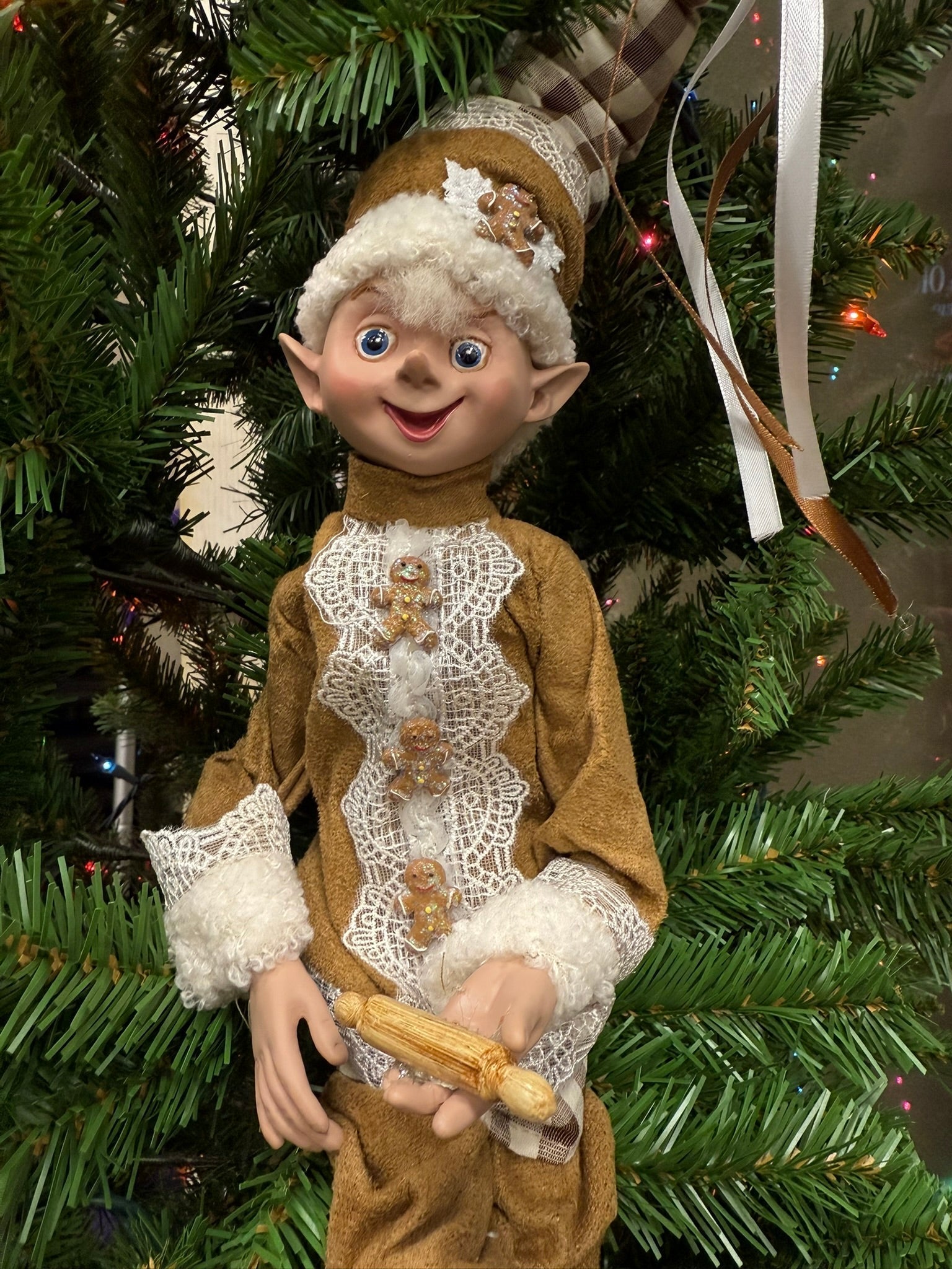 Decorative elf figurine in a brown outfit with lace details, holding a rolling pin, against a Christmas tree background.